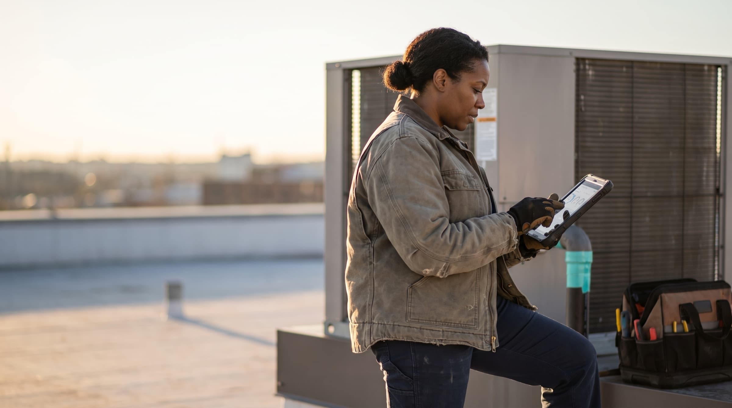 An HVAC contractor reviews a job on her tablet beside a rooftop unit at golden hour
